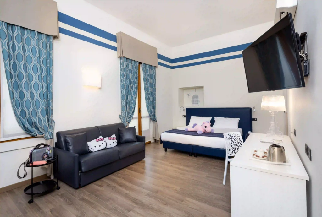 Nell'immagine, la camera Superior dell'Hotel Genio: si vede il letto matrimoniale su cui è poggiato un peluche. Sulla sinistra, un divano con due cuscini di Hello Kitty e, di fronte, la TV appesa alla parete e un tavolo su cui è poggiato un vassoio con bollitore e tazze.